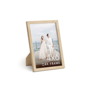Puede incluir: Un marco de fotos de madera clara con una abertura de 10,2 x 15,2 cm. El marco está de pie sobre una superficie blanca. El marco tiene una foto de una pareja caminando por un muelle de madera. El texto "4x6 OAK FRAME" está impreso en el marco.