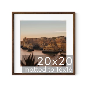 May include: A framed photograph of a rocky coastline with a sunset sky. The image is matted to 16x16 inches and framed in a brown wood frame. The text "20x20 matted to 16x16" is printed below the image.