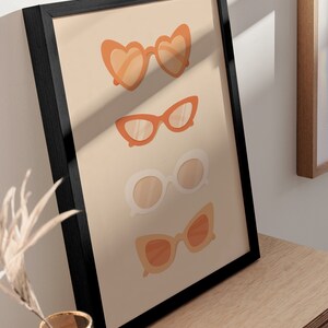 May include: A framed print with a beige background featuring four pairs of sunglasses in different styles. The sunglasses are all in shades of orange and white.
