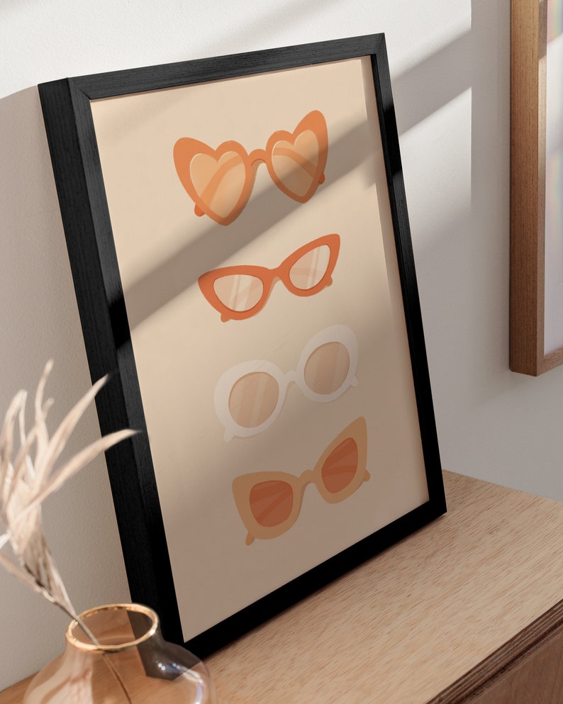 May include: A framed print with a beige background featuring four pairs of sunglasses in different styles. The sunglasses are all in shades of orange and white.