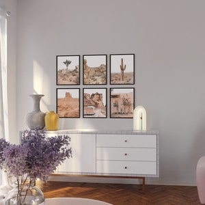 Pu&ograve; includere: Una cassettiera bianca con tre cassetti e pomelli dorati si trova in un soggiorno con un pavimento in legno chiaro. Una lampada bianca con un arco dorato si trova sopra la cassettiera. Sei stampe incorniciate di paesaggi desertici sono appese al muro sopra la cassettiera.