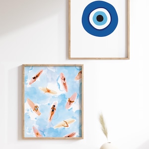 May include: Two framed prints on a white wall. The top print features a blue evil eye symbol on a white background. The bottom print features a watercolor illustration of people on surfboards in the ocean.