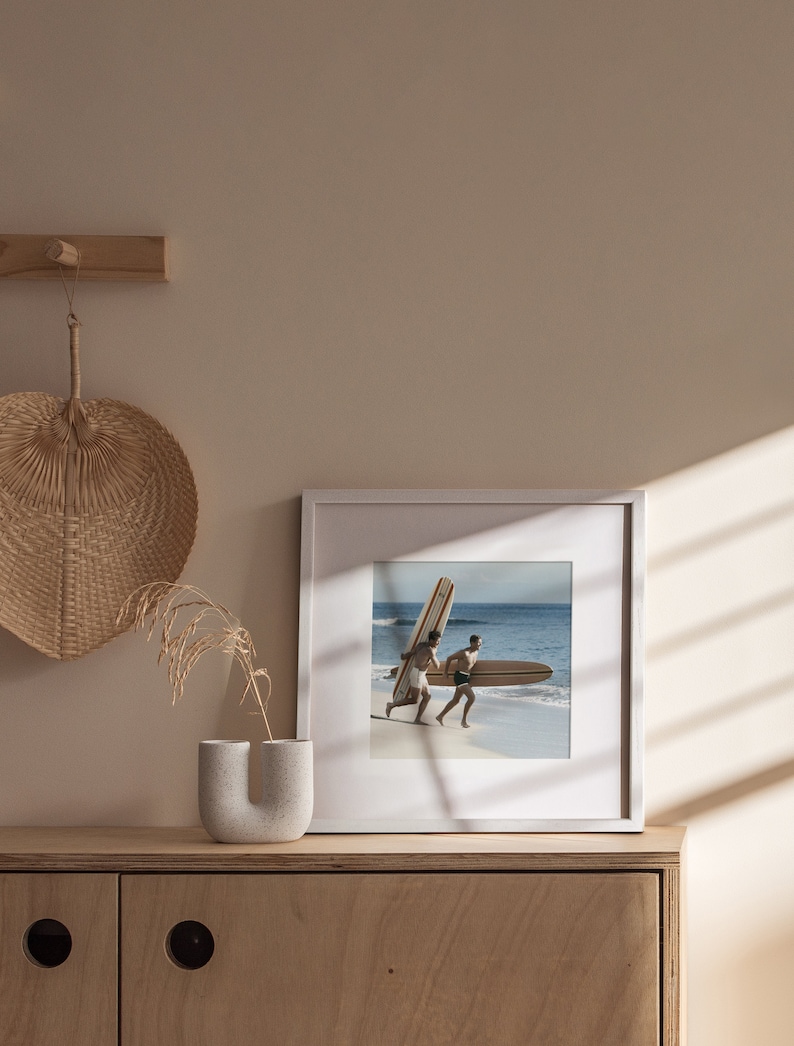 Op de afbeelding: Een wit ingelijste foto van twee mensen die op een strand rennen met surfplanken. De foto hangt aan een licht houten wandplank met een geweven waaier en een vaas met droog gras.