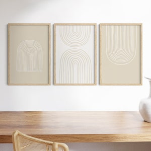 May include: Three framed art prints with abstract arch designs. The prints feature beige and white colors, with the arches in a minimalist style. The frames are made of light-colored wood. The artwork is displayed on a white wall above a wooden desk.