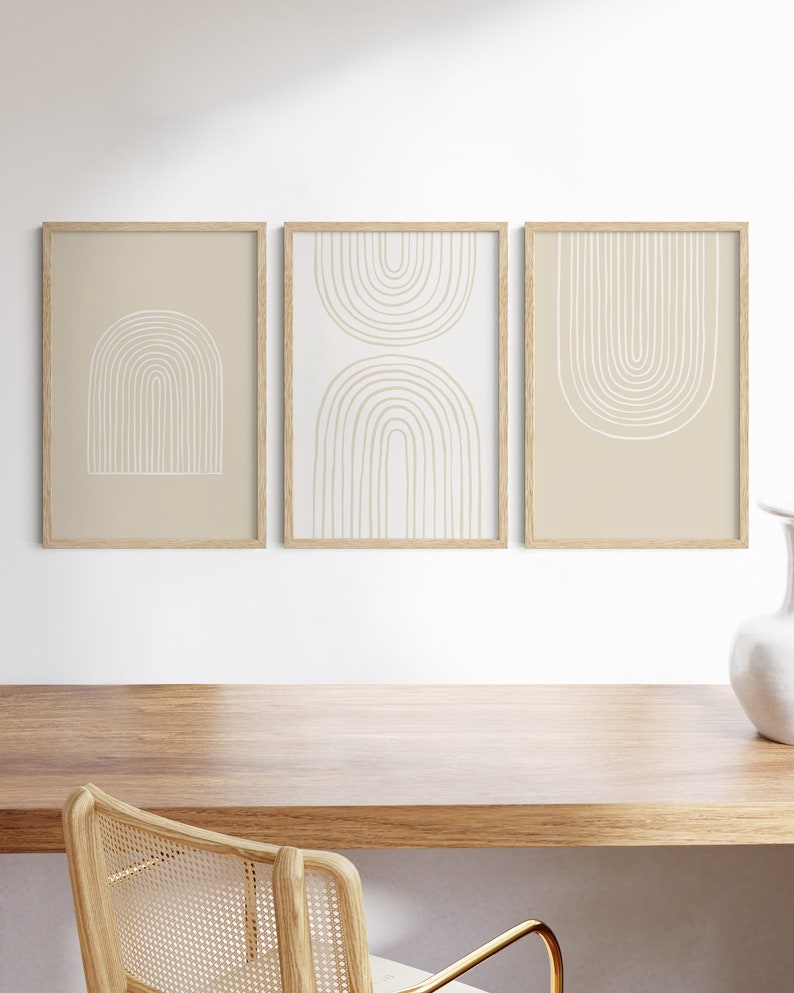May include: Three framed art prints with abstract arch designs. The prints feature beige and white colors, with the arches in a minimalist style. The frames are made of light-colored wood. The artwork is displayed on a white wall above a wooden desk.