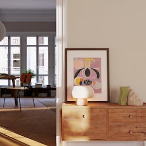 May include: A wooden dresser with two drawers, a white ceramic table lamp with a mushroom-shaped shade, and a framed abstract art print with pink, yellow, and black colors. The dresser is in a room with a large window and a wooden floor.