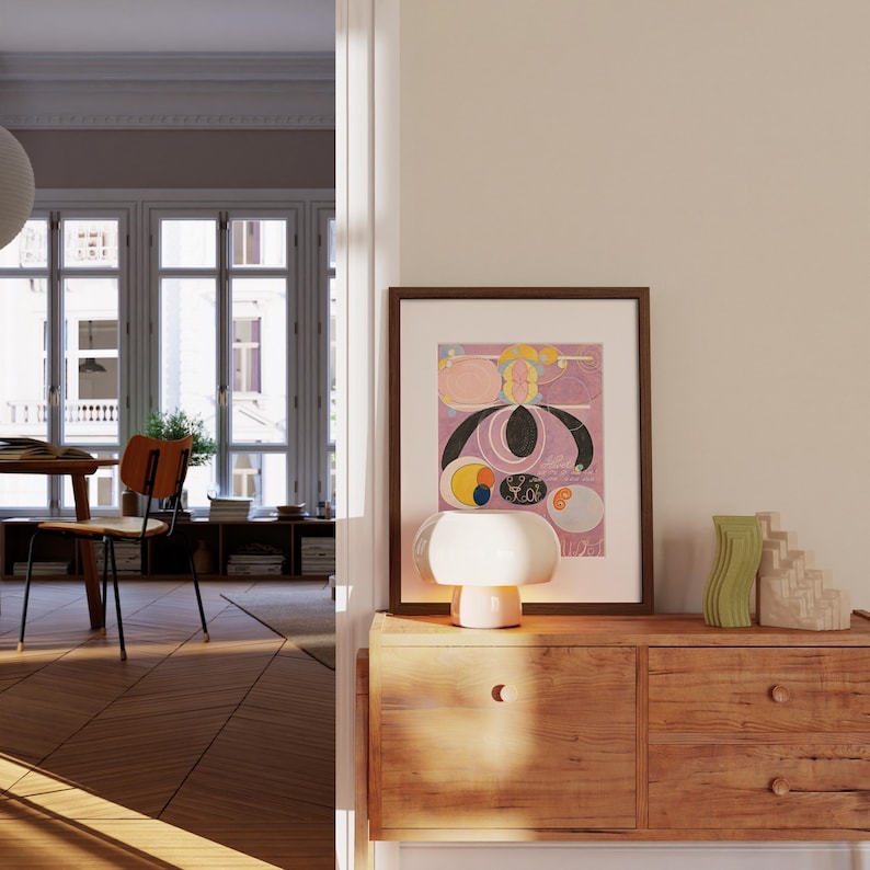 May include: A wooden dresser with two drawers, a white ceramic table lamp with a mushroom-shaped shade, and a framed abstract art print with pink, yellow, and black colors. The dresser is in a room with a large window and a wooden floor.