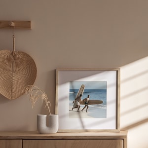 May include: A framed photograph of two people running on a beach, each carrying a surfboard. The frame is light wood, and the image is set against a neutral wall. A decorative fan and vase are on a wooden cabinet.