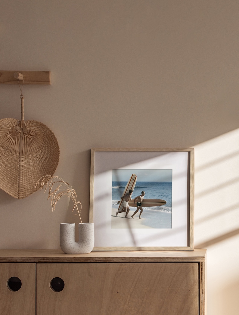 May include: A framed photograph of two people running on a beach, each carrying a surfboard. The frame is light wood, and the image is set against a neutral wall. A decorative fan and vase are on a wooden cabinet.