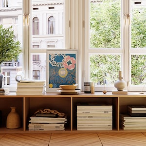 May include: A wooden shelf unit with three shelves, each filled with books and magazines. A framed print with a blue background and colorful abstract shapes hangs on the wall above the shelf. A vase and a bowl are on the shelf.