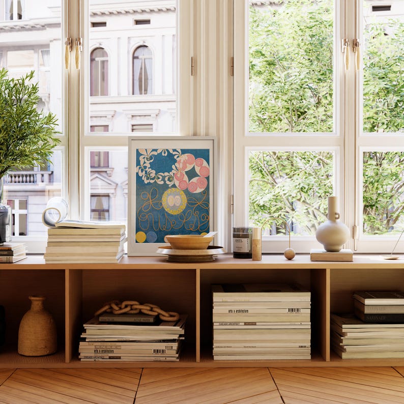 May include: A wooden shelf unit with three shelves, each filled with books and magazines. A framed print with a blue background and colorful abstract shapes hangs on the wall above the shelf. A vase and a bowl are on the shelf.