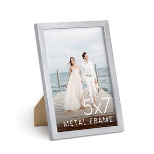 May include: A silver metal picture frame with a white mat, holding a photo of a couple walking on a dock. The frame is 5x7 inches and has the text "5x7 Metal Frame" printed on the bottom.