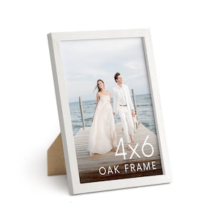 May include: A white oak frame with a 4x6 inch photo. The frame has a simple design and a stand on the back. The photo shows a couple walking on a wooden dock near the water.