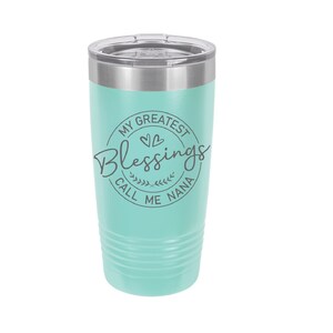 May include: A turquoise stainless steel tumbler with a silver lid. The tumbler has a white circle with the text "My Greatest Blessings Call Me Nana" and two small hearts.