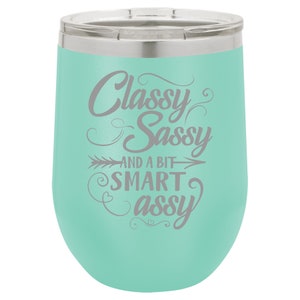 May include: A turquoise stainless steel wine tumbler with a silver lid. The tumbler has a gray inscription that reads "Classy Sassy and a bit Smart Assy".