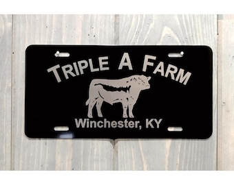 May include: A rectangular black license plate with rounded corners featuring the silver text "TRIPLE A FARM" arched above a silver silhouette of a bull, with the text "Winchester, KY" printed in silver below the bull and four mounting slots.