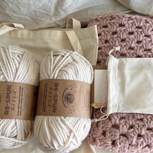 Puede incluir: Un kit de manualidades con dos madejas de hilo blanco roto con etiquetas marrones, un artículo de ganchillo rosa claro, una bolsa de lona y una pequeña bolsa con cordón. Las etiquetas del hilo dicen "RE-SPUN" y "100% RECYCLED POLYESTER".