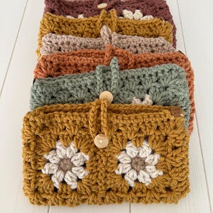 May include: A stack of crocheted pouches with floral designs in various colours, including mustard yellow, green, orange, beige, and burgundy. Each pouch has a wooden button closure and a crocheted strap.