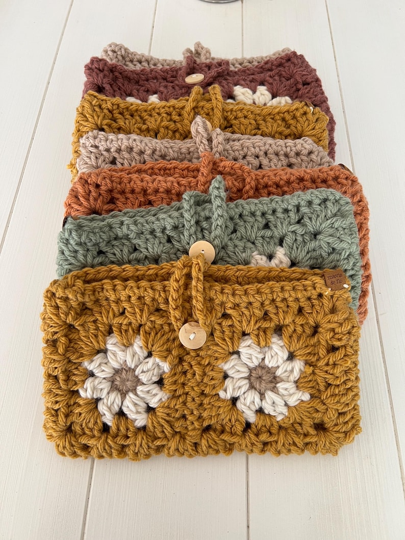 May include: A stack of crocheted pouches with floral designs in various colours, including mustard yellow, green, orange, beige, and burgundy. Each pouch has a wooden button closure and a crocheted strap.