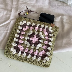 May include: A square, crocheted pouch in a granny square pattern. The pouch features a colour scheme of olive green, pink, brown, and white. A black smartphone is partially visible inside. The zipper pull is a leather cord with beads.