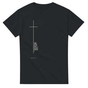 Könnte beinhalten: Ein schwarzes T-Shirt mit kurzen Ärmeln und einer vertikalen weißen Grafik, die ein Kreuz bildet. Das Wort "still." ist in weißen Kleinbuchstaben entlang der vertikalen Linie gedruckt, darunter befindet sich der Text "PSALM 46".