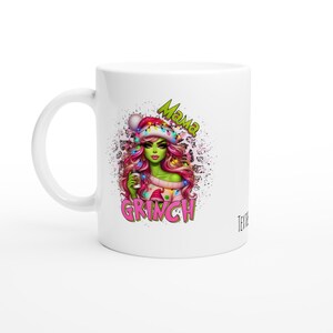 Könnte beinhalten: Ein weißer Keramikbecher mit einer grafischen Illustration einer grünhäutigen Figur mit langem rosa Haar, die eine Weihnachtsmütze mit bunten Lichterketten trägt. Der Text Mama GRINCH ist in grünen und rosa Buchstaben aufgedruckt.