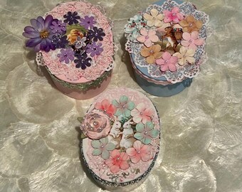 May include: Three oval-shaped decorative boxes with floral embellishments. The boxes are pink, blue, and white with lace trim. Each box features a different floral design with a central image of a rabbit or a bird.