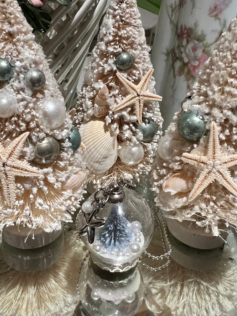 May include: Coastal-themed Christmas tree decorations. Three small, snow-covered bottle brush trees are adorned with seashells, starfish, and silver ornaments. A clear glass ornament with a miniature tree and pearls hangs in front.