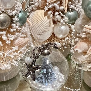 May include: Close-up of decorative Christmas ornaments with a coastal theme. The ornaments feature seashells, starfish, and small glass globes with miniature trees and pearl accents. The color palette includes white, silver, and touches of blue.
