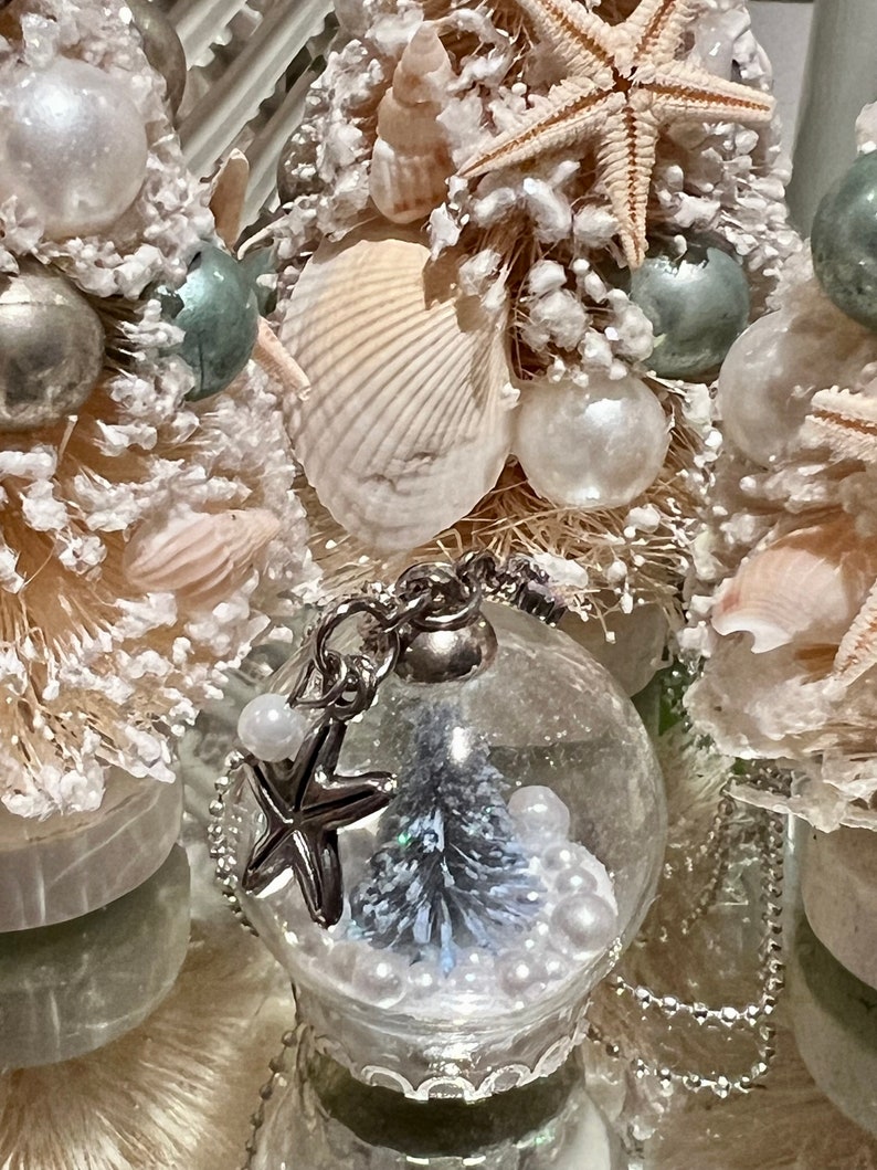 May include: Close-up of decorative Christmas ornaments with a coastal theme. The ornaments feature seashells, starfish, and small glass globes with miniature trees and pearl accents. The color palette includes white, silver, and touches of blue.