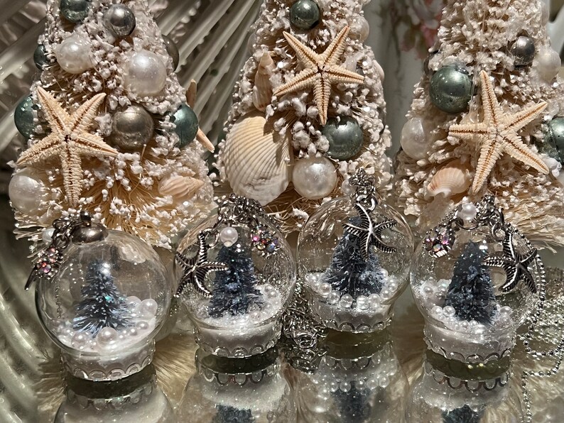 May include: Coastal-themed Christmas ornaments and miniature trees. The trees are white with silver and teal accents, seashells, and starfish. Clear glass ball ornaments contain miniature trees, pearls, and starfish. A festive, beachy holiday theme.