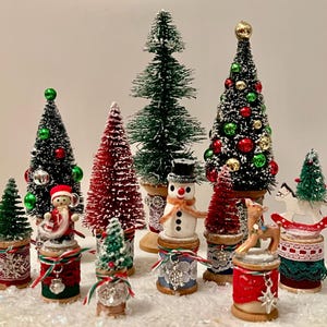 Puede incluir: Una colección de miniaturas navideñas, que incluye árboles de Navidad, muñecos de nieve y renos. Los árboles están adornados con adornos y se asientan sobre carretes de madera adornados con cintas y encajes. La paleta de colores incluye verde, rojo y blanco.