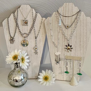 May include: Three silver necklaces with daisy-themed pendants displayed on a white wooden stand. The necklaces feature a variety of chain styles, including chunky, delicate, and layered. The pendants include a square with a daisy design, a round pendant with a daisy design, and a daisy charm with a butterfly.