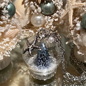 May include: A clear glass globe pendant necklace with a miniature blue Christmas tree inside, surrounded by white beads. The pendant is adorned with a silver starfish charm and hangs on a silver chain. The background features decorative seashell Christmas trees.