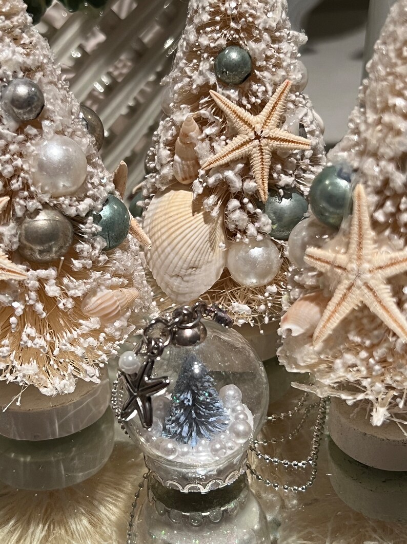 May include: Coastal-themed Christmas decorations featuring miniature bottle brush trees adorned with seashells, starfish, and pearl-like ornaments. A clear glass snow globe with a miniature tree and pearls is in the foreground. The trees are on a reflective surface.