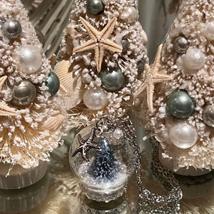 May include: Three miniature Christmas trees decorated with seashells, pearls, and starfish. A silver necklace with a glass globe pendant containing a miniature tree and starfish charm is in the foreground. The trees are on a reflective surface.