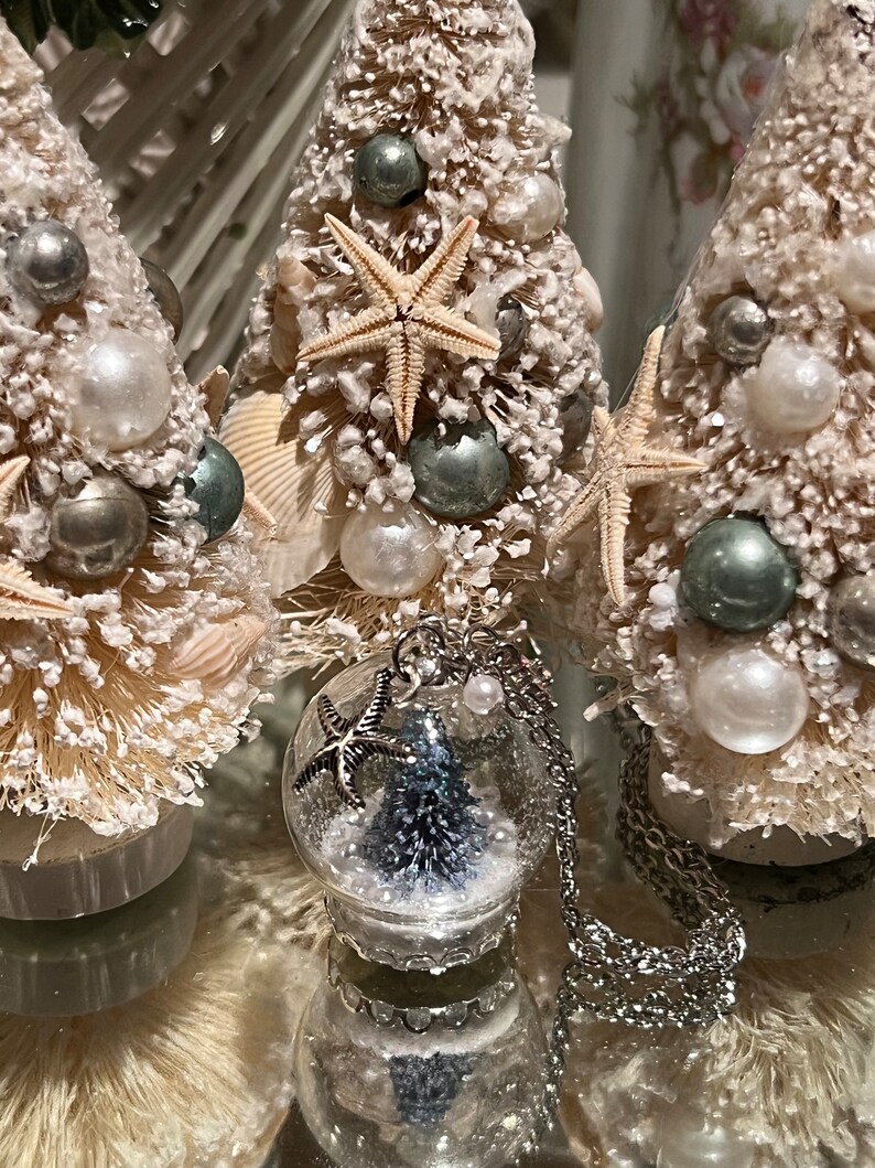 May include: Three miniature Christmas trees decorated with seashells, pearls, and starfish. A silver necklace with a glass globe pendant containing a miniature tree and starfish charm is in the foreground. The trees are on a reflective surface.