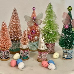May include: A collection of decorative miniature bottle brush Christmas trees in various colors, including pink, green, and orange. Some trees are adorned with butterflies, beads, and ornaments. Easter eggs and a small white bunny are also present.