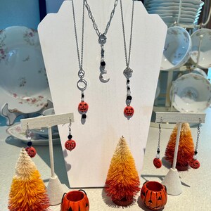 Peut inclure: Un ensemble de bijoux sur le thème d'Halloween comprenant des boucles d'oreilles et des colliers en forme de citrouille avec des chaînes argentées, des perles noires et des breloques en forme de citrouille. Les bijoux sont présentés sur un présentoir blanc avec de petits arbres orange et blancs.
