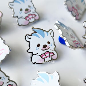 May include: A collection of enamel pins featuring a cartoon bear design. The bears are white with blue accents on their hair and paws, and hold a pink seashell. The pins have a shiny, metallic finish.