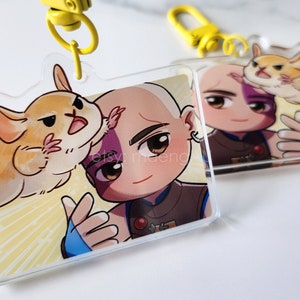 May include: A clear acrylic keychain featuring a cartoon character with a shaved head, wearing a blue and brown outfit, holding a small, light brown hamster. The background is a yellow and white pattern.