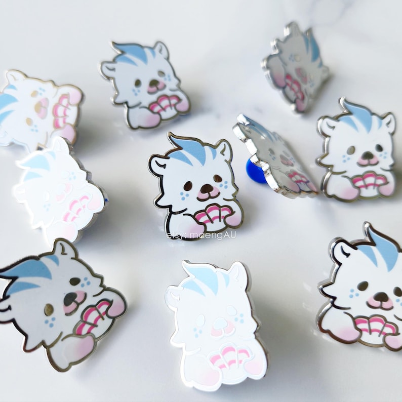 May include: A collection of enamel pins featuring a cartoon bear design. The bears are white with blue accents on their hair and paws, and hold a pink seashell. The pins have a shiny, metallic finish.