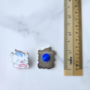 May include: Two enamel pins. One pin is a white tiger with blue stripes and pink details. The other pin is a brown shape with a blue pin back. A ruler is next to the pins. The text "etsy: maengAU" is visible.