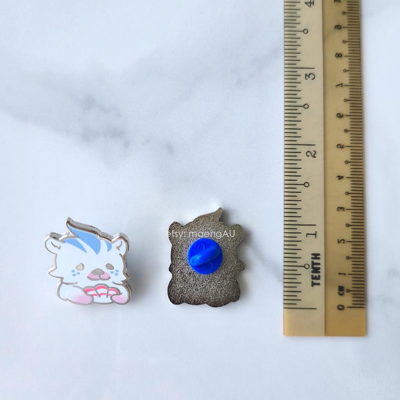May include: Two enamel pins. One pin is a white tiger with blue stripes and pink details. The other pin is a brown shape with a blue pin back. A ruler is next to the pins. The text "etsy: maengAU" is visible.