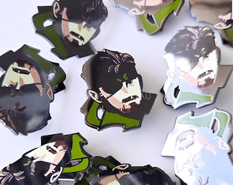 May include: Enamel pins featuring a stylized portrait of a character with black hair, a beard, and a green and silver outline. The pins have a glossy finish and are scattered across a white surface.