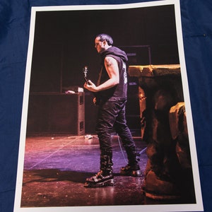 Puede incluir: Un hombre con una camisa negra y pantalones negros toca un bajo en un escenario. Lleva botas negras con hebillas. El escenario está iluminado con luces moradas y rojas.