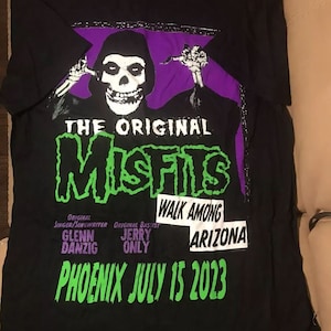 May include: Black t-shirt with a green and white graphic of The Misfits band logo. The text "The Original Misfits" is above the logo. The text "Walk Among Arizona" is below the logo. The text "Phoenix July 15 2023" is at the bottom of the graphic.