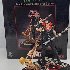 Puede incluir: Una figura de la serie Misfits Rock Iconz Collector. La figura es de un músico con un atuendo negro con un cuello rojo, sosteniendo una guitarra y un esqueleto. La caja tiene el logotipo de Misfits en verde.