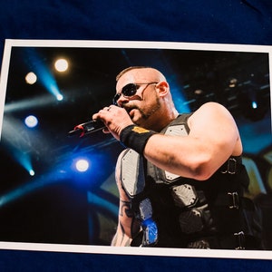 May include: A bald man with a beard and sunglasses is singing into a microphone. He is wearing a black vest with a silver and black pattern. The background is dark with blue and white lights.