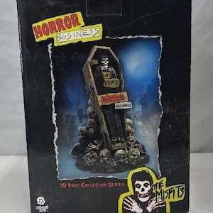 Puede incluir: Caja de la serie de coleccionista de vinilo 3D con una figura de un esqueleto en un ataúd, rodeado de calaveras. La caja tiene las palabras "HORROR BUSINESS" y "THE MISFITS" en amarillo y negro. El fondo muestra una escena de cementerio.